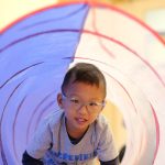 A young boy wearing glasses and a gray shirt crawls through a play tunnel, smiling at the camera. The tunnel is light purple with red stripes and is indoors.