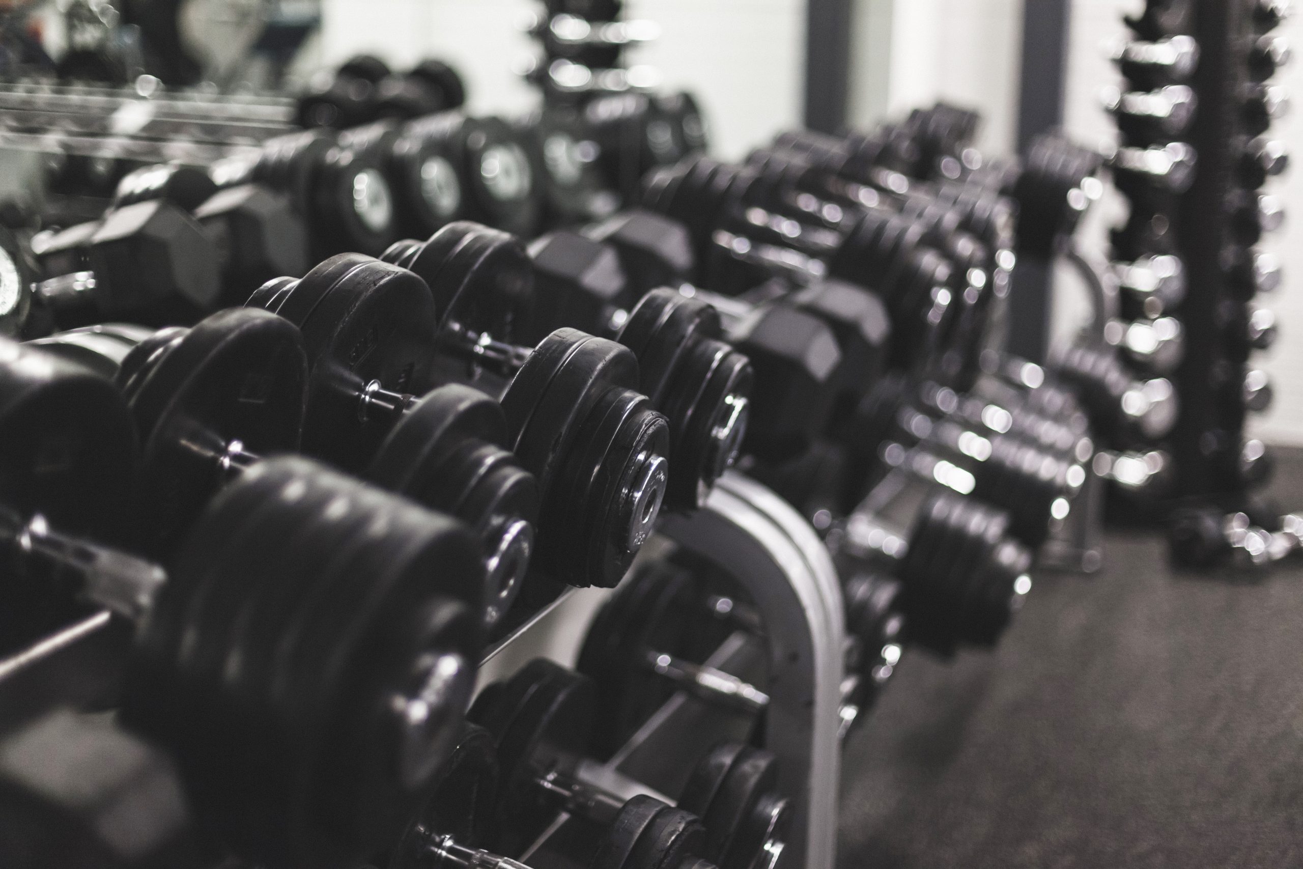 Rows of black dumbbells arranged on a metal rack in a gym, with more weights and gym equipment visible in the blurred background.