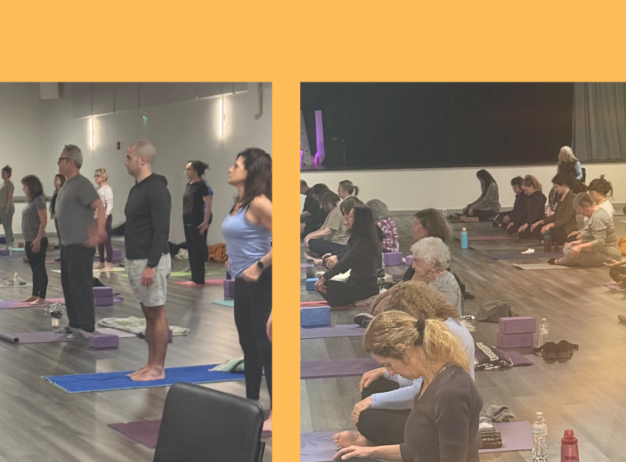 A group of people in an indoor studio participate in a yoga class, standing or sitting on mats while facing the instructor. The room is well-lit, and yoga props are placed on the floor near participants.