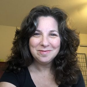 A woman with wavy brown hair wearing a black shirt smiles at the camera in a softly lit indoor setting with a beige wall and part of a folding screen in the background.