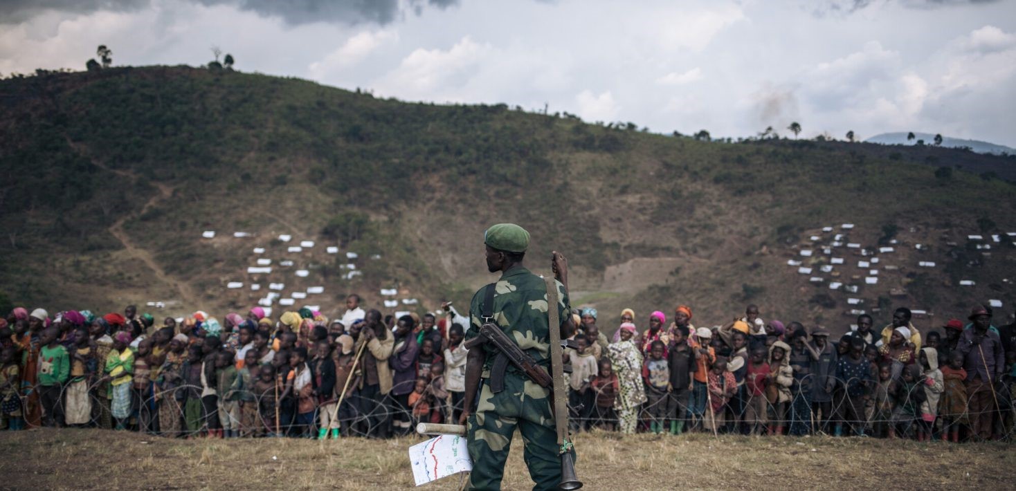 A soldier with a rifle stands facing a large group of people behind barbed wire, set against a backdrop of green hills dotted with small white shelters under a cloudy sky.