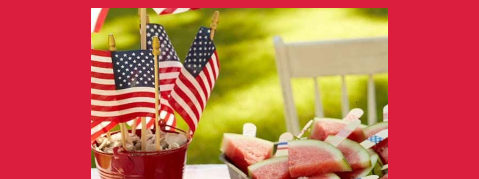 Small American flags in a red bucket next to a plate of watermelon slices with popsicle sticks, set outdoors on a sunny day with a white chair and green grass in the background.