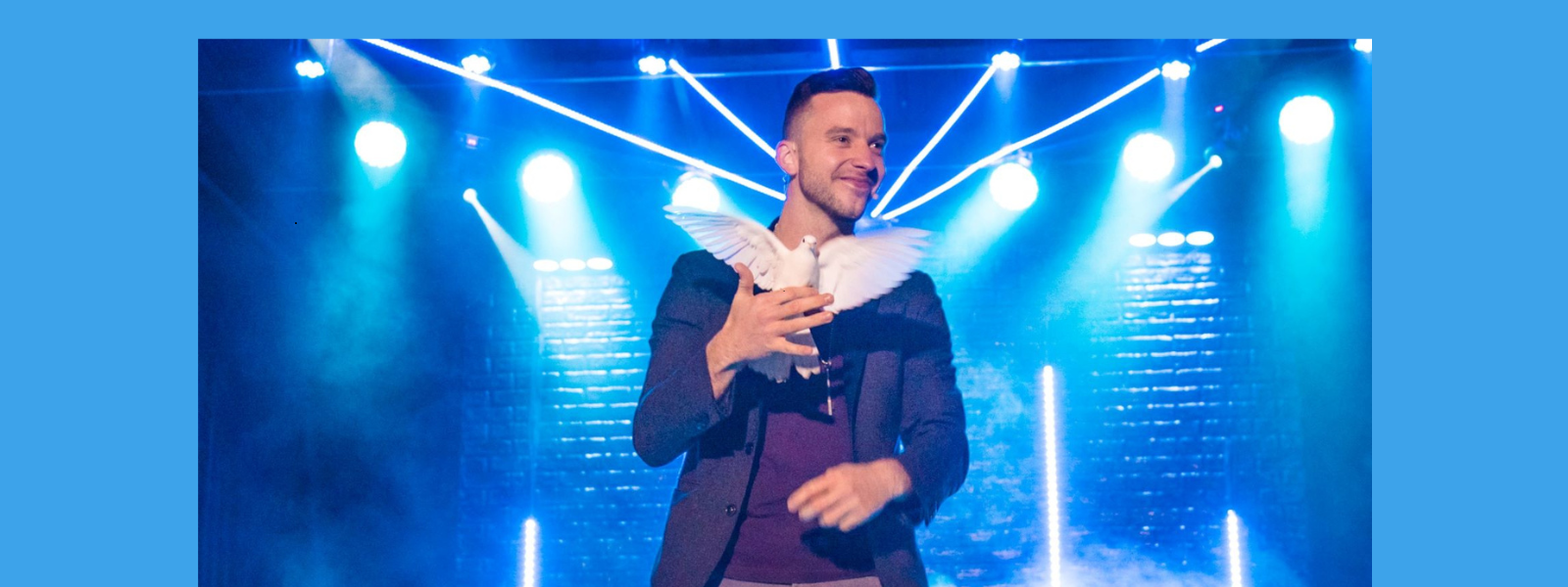 A magician in a spotlight holds a white dove, smiling on stage. Blue lights and smoke create a mystical atmosphere with a sense of wonder.