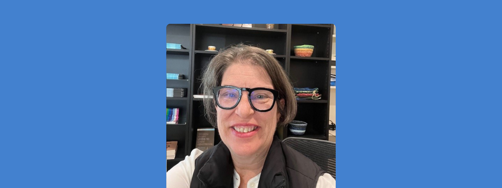 A person with glasses smiles warmly in an office. Behind are shelves with books and colorful decorations, creating a welcoming atmosphere.