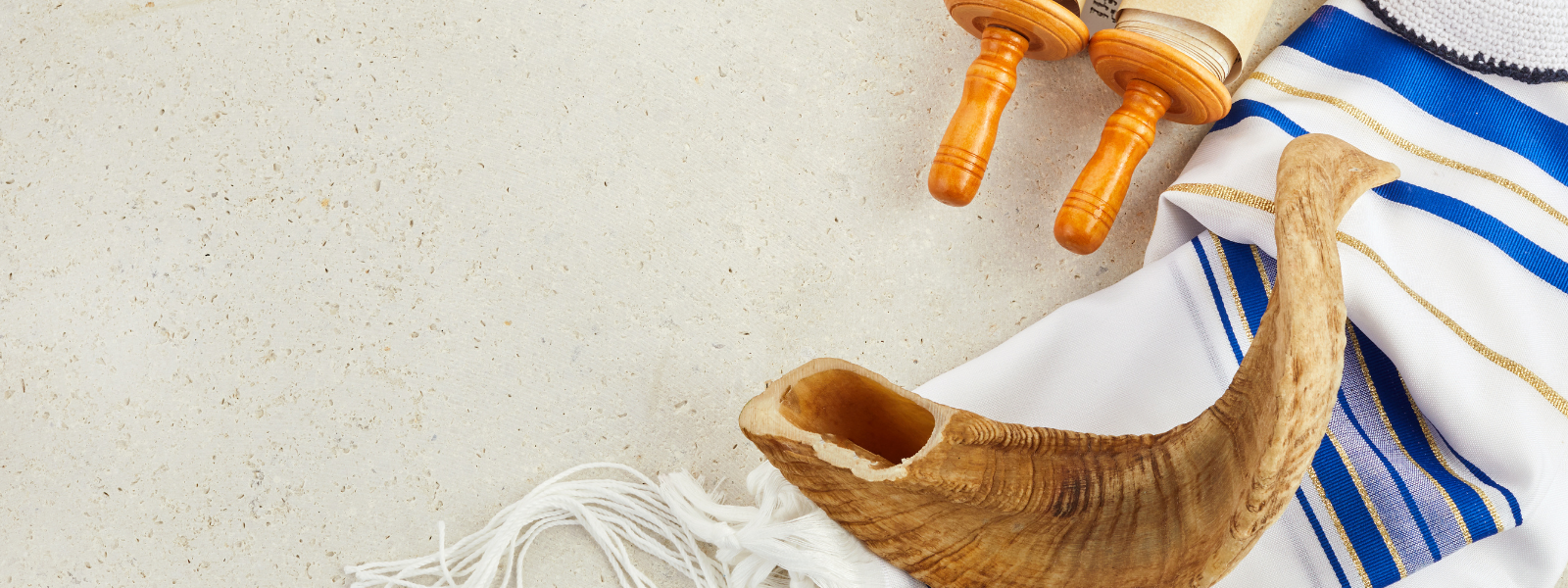 A shofar, prayer shawl, and Torah scroll with ornate handles rest on a light textured surface. The mood is reflective and reverent, signifying Jewish traditions.