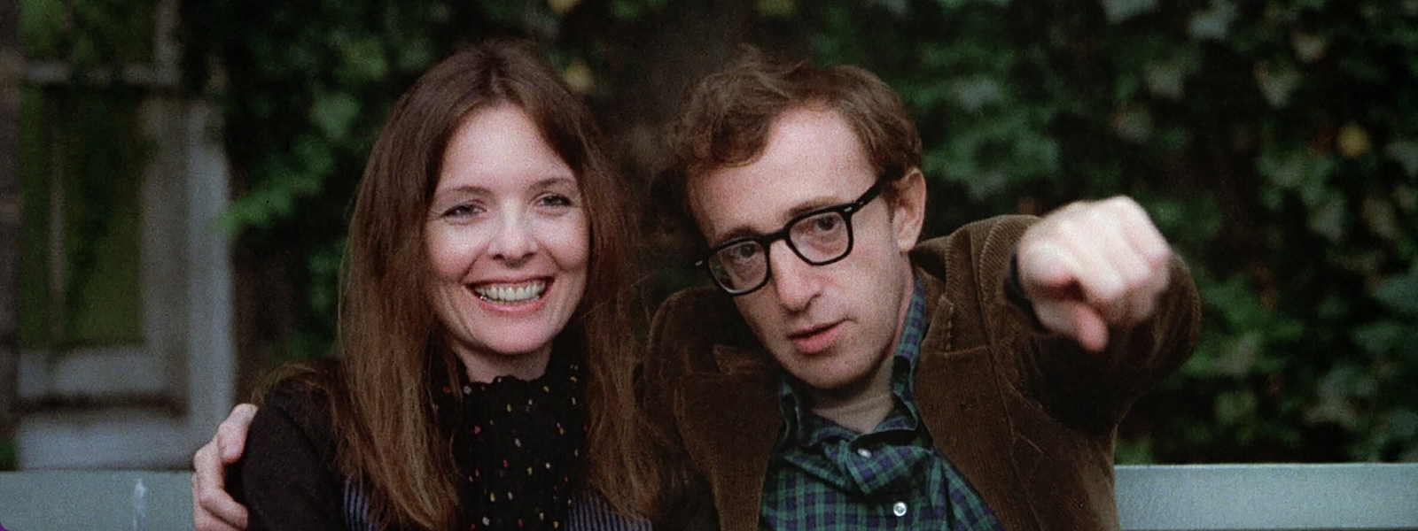 A woman and a man sit on a bench, smiling. The man points forward. They are outdoors with ivy-covered walls behind them, creating a relaxed, happy atmosphere.