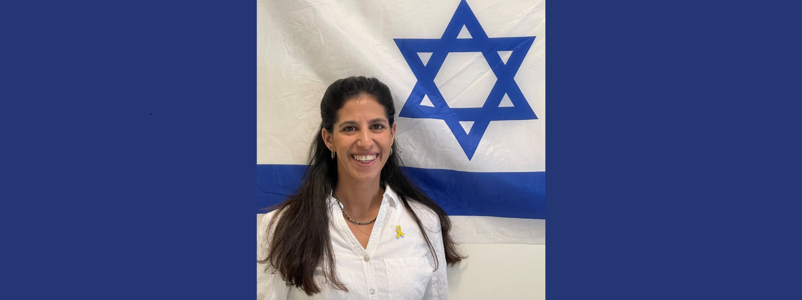 A woman in a white shirt, smiling, stands in front of an Israeli flag, featuring a blue Star of David. The mood is positive and proud.