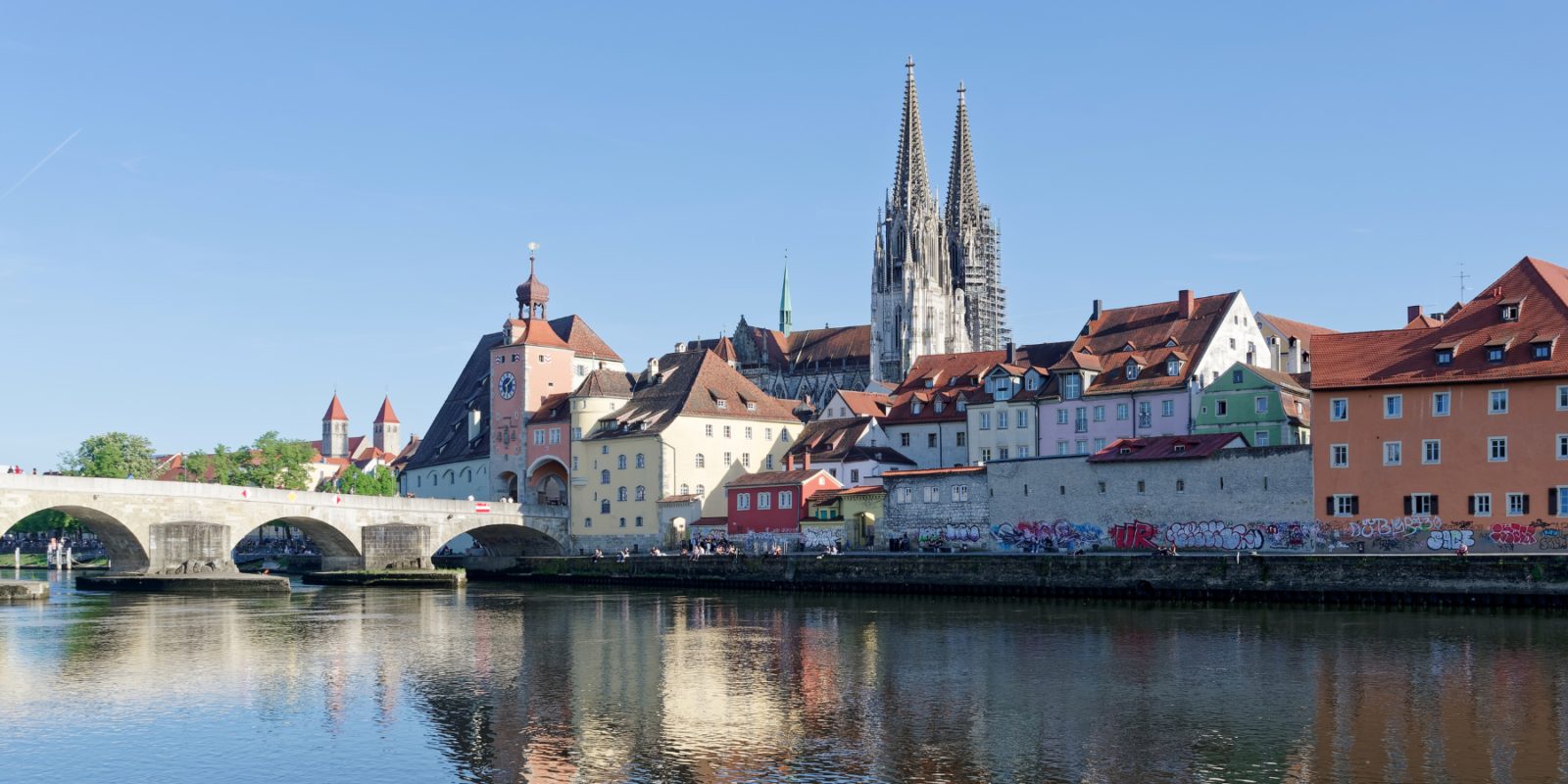Scenic view of a historic European town with colorful buildings along a river. A stone bridge crosses the water, and twin-spired cathedral towers rise in the background under a clear blue sky.