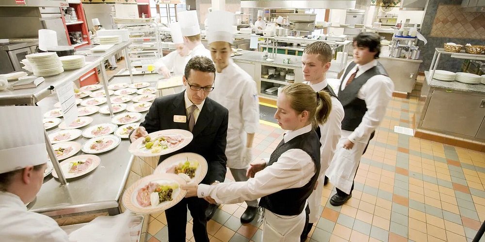 Restaurant kitchen scene with chefs and waitstaff in uniforms, preparing and serving dishes. The environment is busy yet orderly, conveying teamwork.