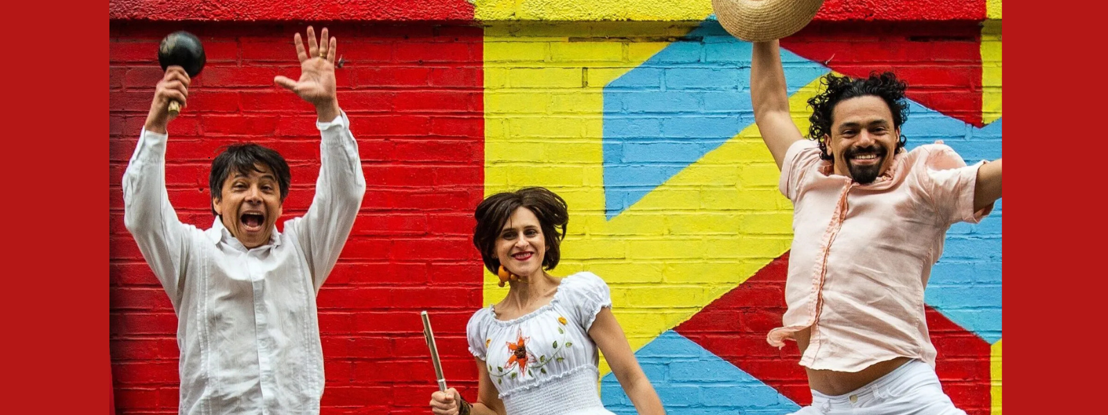Three people in vibrant clothing jump joyfully in front of a colorful mural with red, yellow, and blue shapes. Two men hold hats, and a woman smiles. Energetic and lively.
