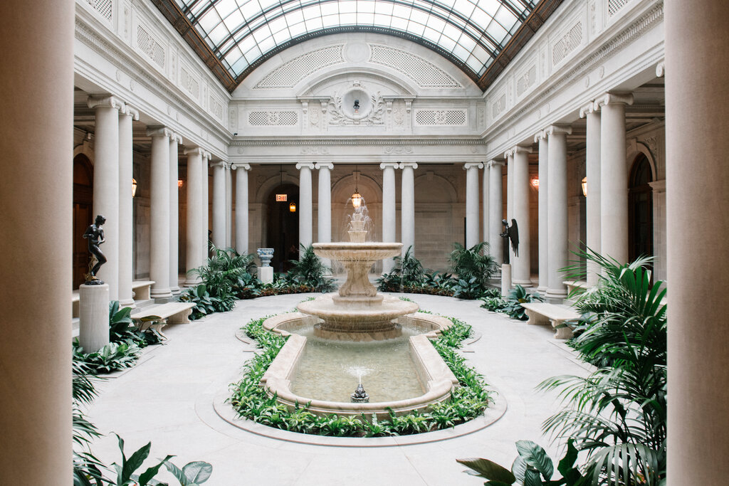 Elegant indoor courtyard with a central fountain, surrounded by plants and classical columns. Soft lighting and serene atmosphere under a glass ceiling.