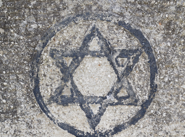 A Star of David is drawn on a textured concrete surface within a circle. The black, weathered lines contrast with the gray, rough background.