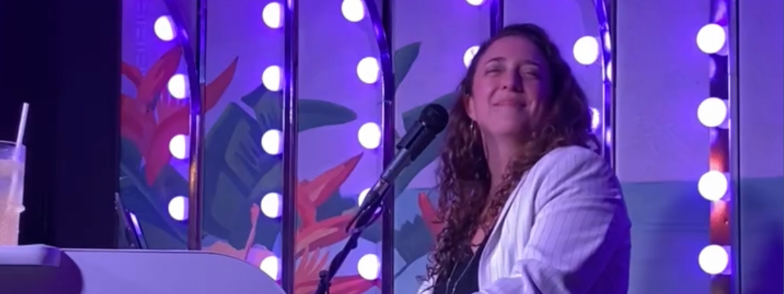 A person in a white jacket smiles while playing piano on a vibrant stage with purple lighting and round bulbs. The scene conveys joy and energy.