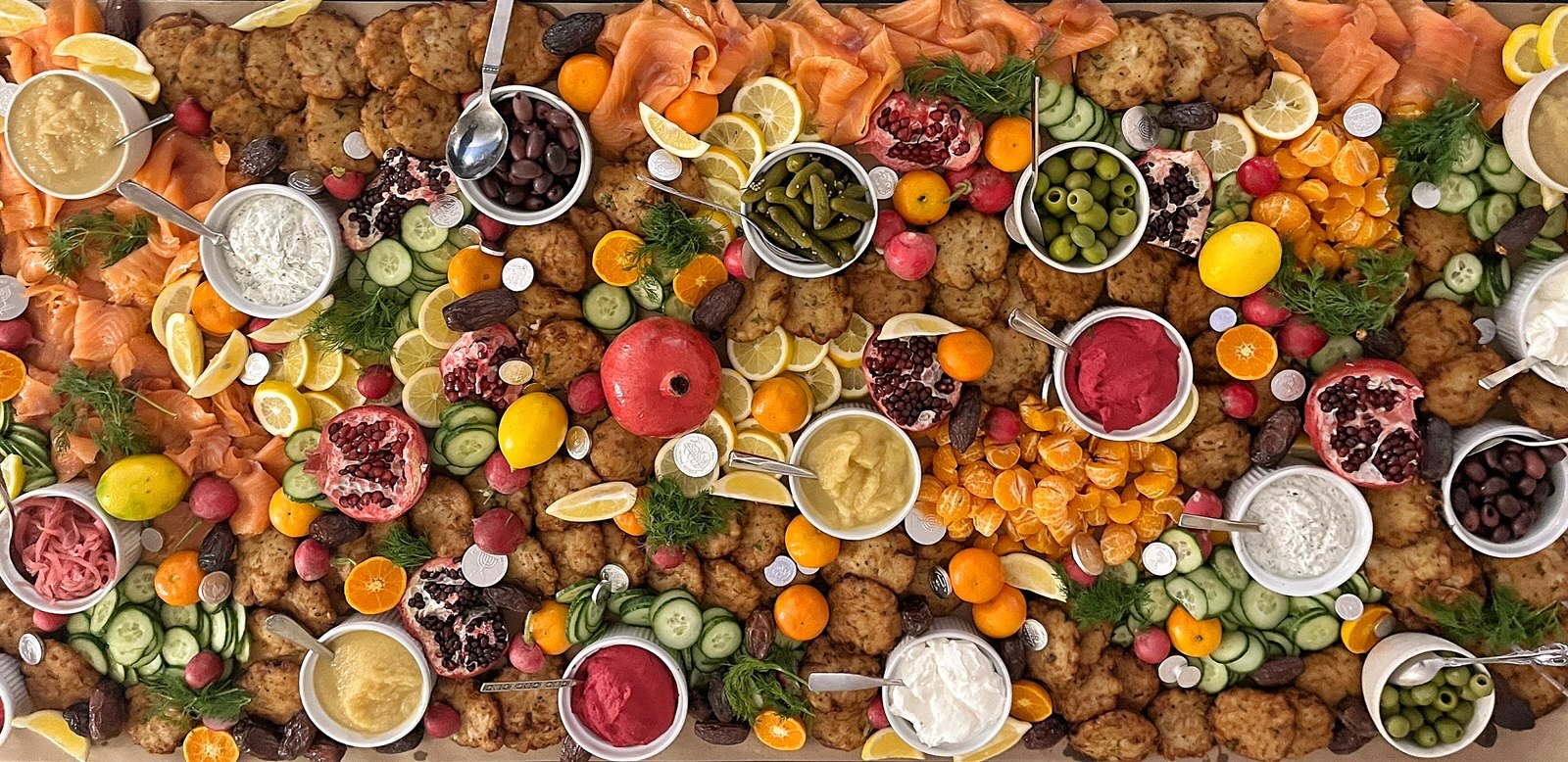 A vibrant spread of diverse foods on a table, including smoked salmon, sliced cucumbers, various dips, citrus slices, and small fruits. The tone is festive and colorful.