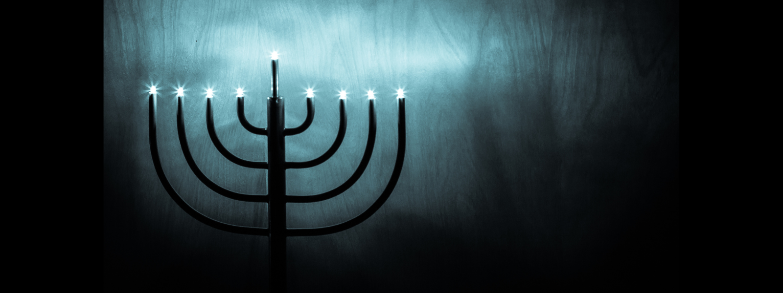 A menorah with seven lit candles stands against a dark, textured background. The soft glow creates an atmosphere of serenity and reflection.