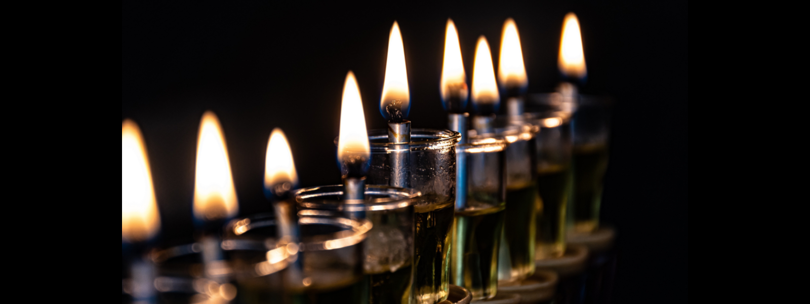 Alt text: "A row of lit oil candles with small flames in glass holders against a dark background, conveying warmth and tranquility."
