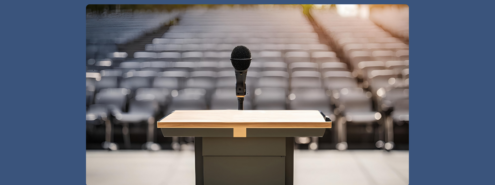 A microphone stands on a podium in an empty auditorium. Rows of blurred gray seats are in the background, conveying a sense of anticipation.